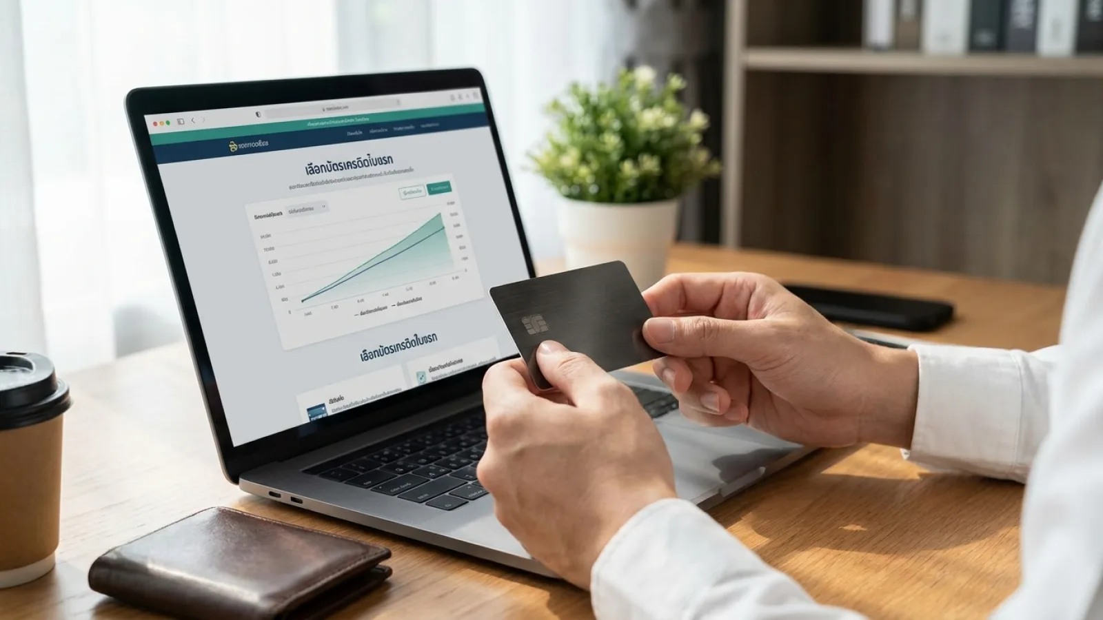 มือของคนกำลังถือบัตรเครดิตใบแรกหน้าแล็ปท็อปบนโต๊ะทำงานที่จัดวางอย่างเป็นระเบียบ แสงธรรมชาติส่องผ่านหน้าต่าง